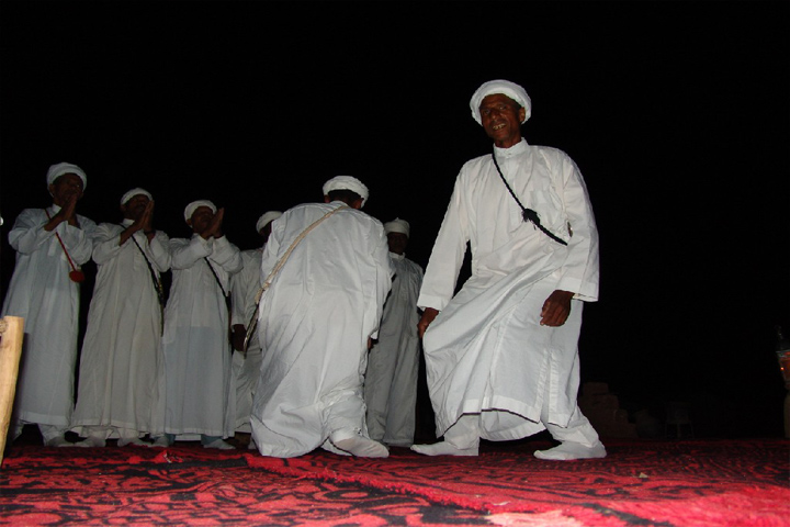 Danseur d'ahwach en djellaba blanche sur un tapis rouge, devant une rangée de chanteurs, lors d'une fête de nuit