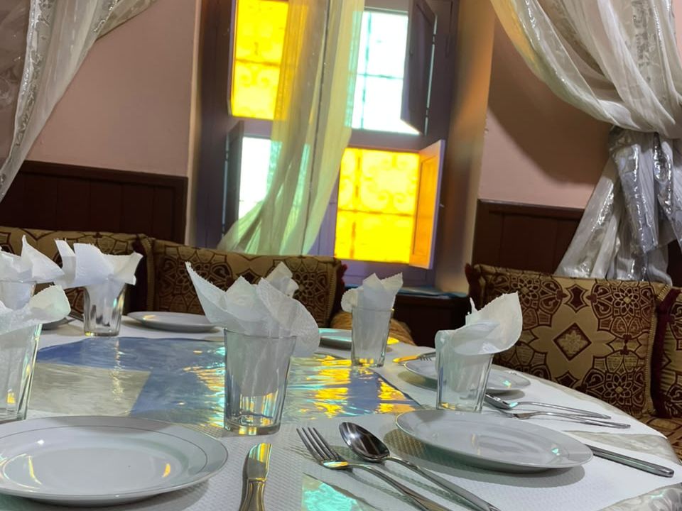 La salle du restaurant de l'auberge à Tagmoute : table dressée, banquettes marocaines et fenêtre aux vitraux colorés