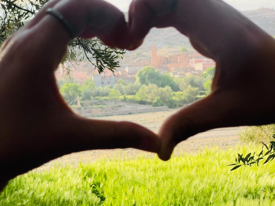 Two hands making a heart shape around the village of Tagmoute, above a green field